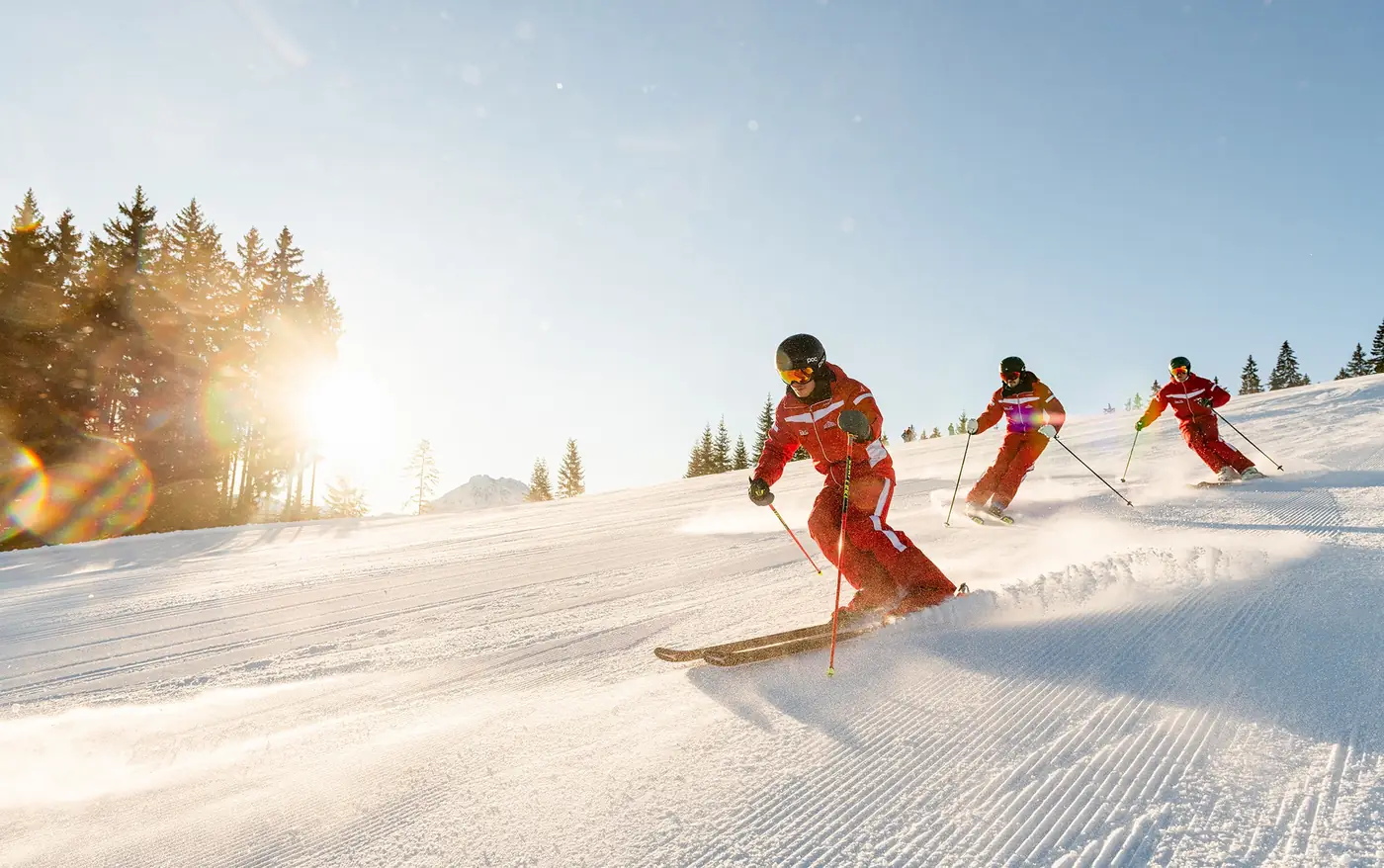 Drei Skilehrer der Skischule Rot-Weiss-Rot carven in der Morgensonne über eine frisch präparierte Piste in Alpendorf