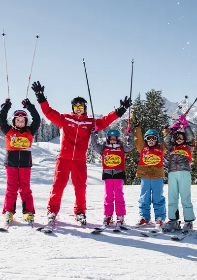 Skilehrer mit fünf jubelnden Kindern im Rennleibchen auf der Piste