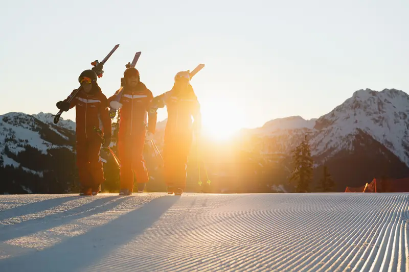 Drei Skilehrer mit geschulterten Ski im Sonnenaufgang auf der Piste