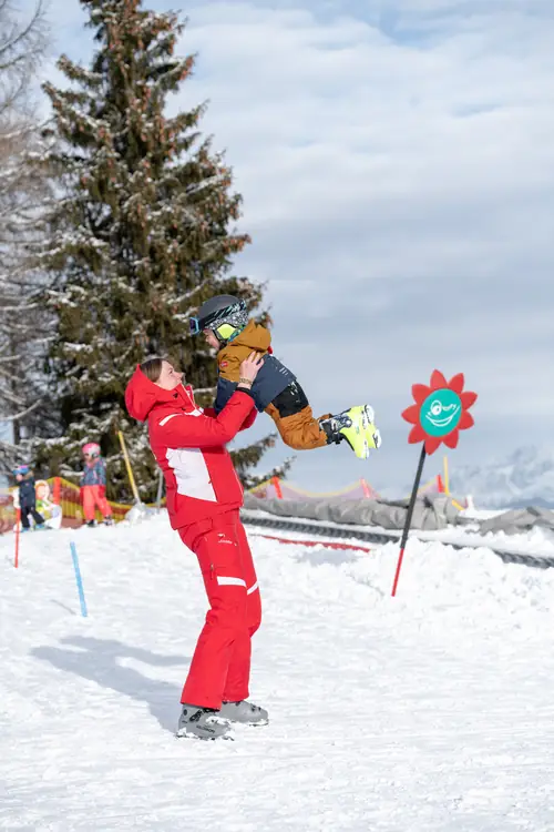 Skilehrerin hebt ein lachendes Kind beim Skispiel in die Luft