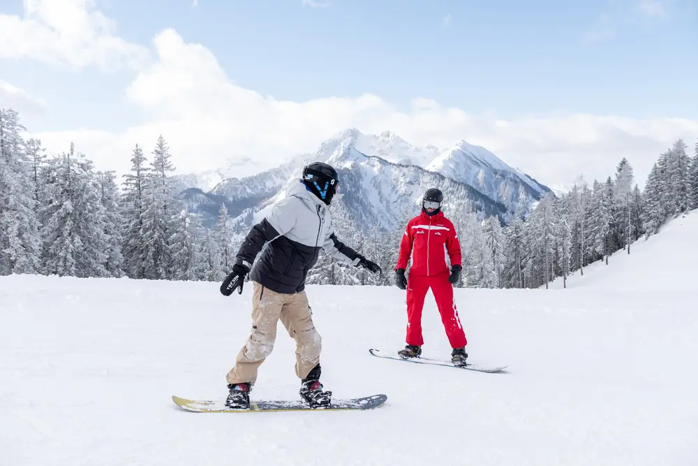Snowboardlehrer begleitet einen Schüler bei der Abfahrt vor verschneiten Bergen